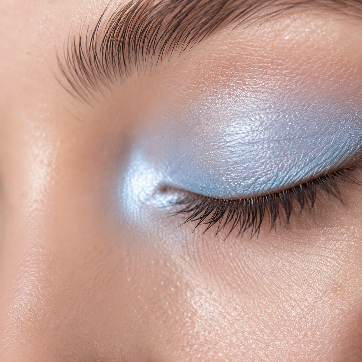 Extreme close-up of a fingertip tapping shimmering icy blue eyeshadow into the inner corner of a closed eye