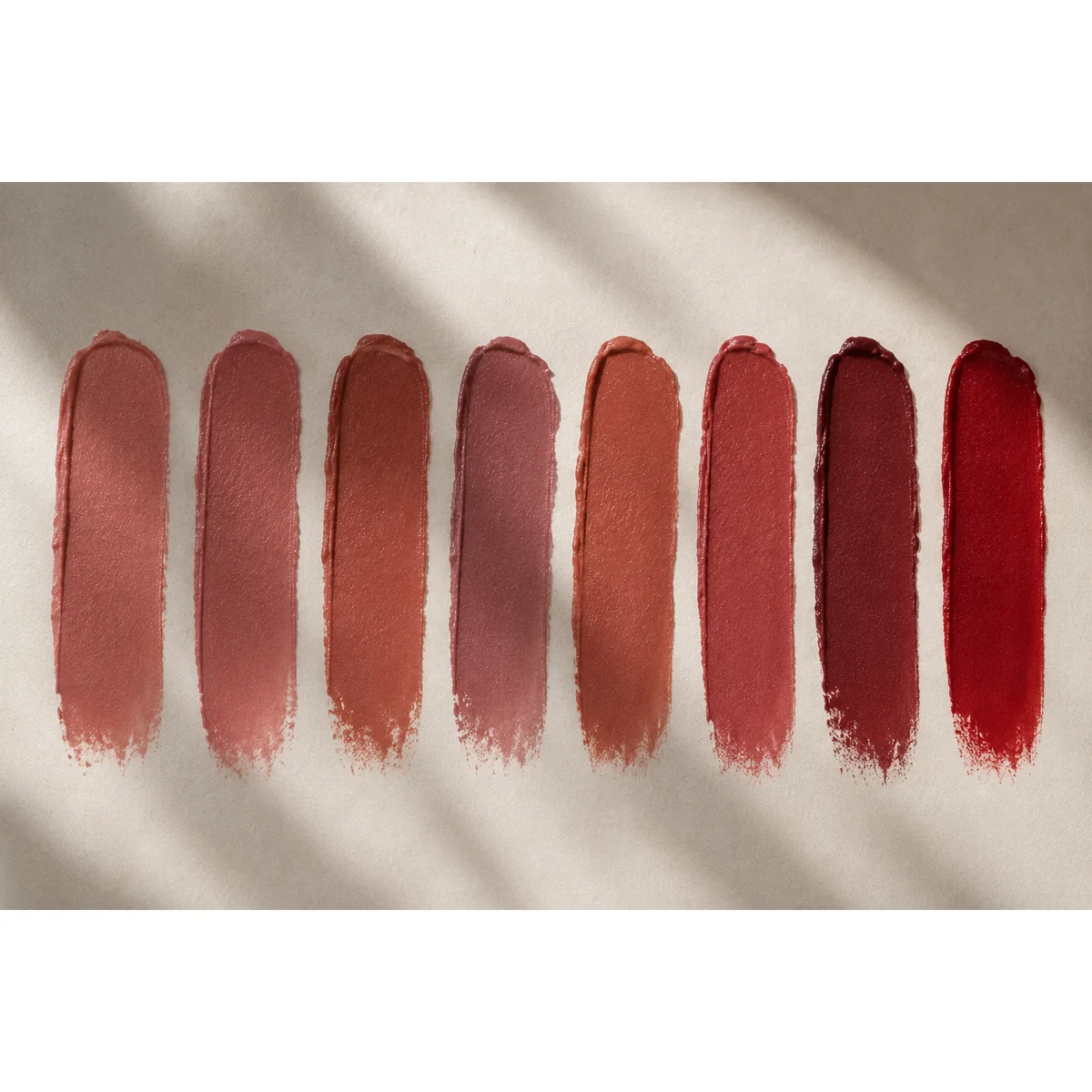 Editorial flatlay of eight lipstick swatches in varying depth and saturation arranged in a row on a neutral surface, soft directional light