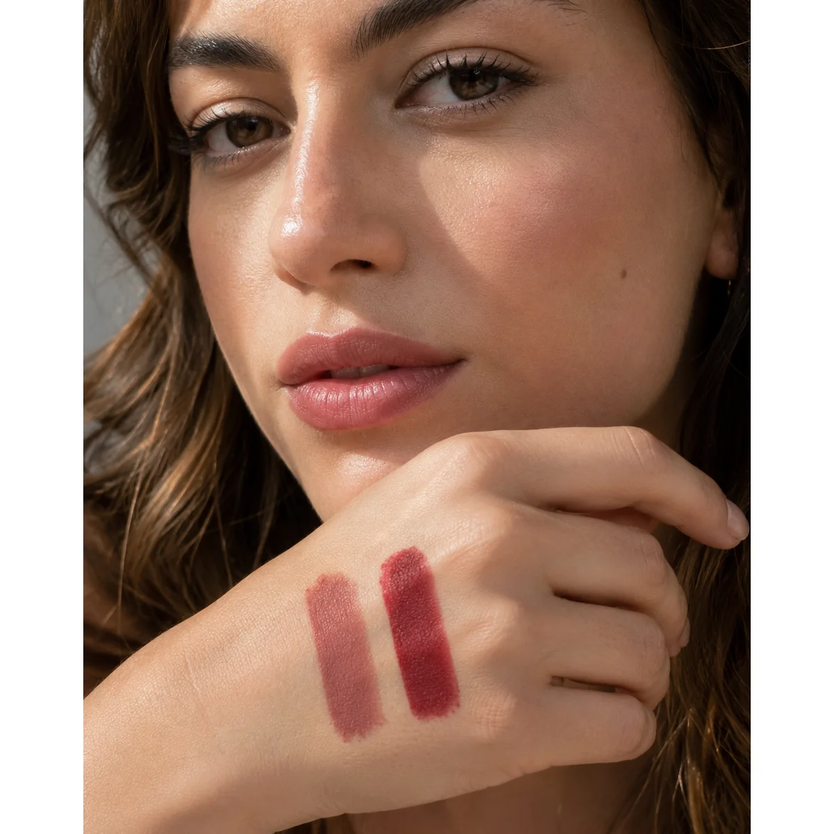 Close-up beauty editorial photo of two lipstick swatches in contrasting intensity, one soft muted rose and one deep saturated red, held near a woman's lips